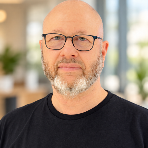 A bald man with glasses and a gray beard, wearing a black shirt against a bright, softly blurred office background.